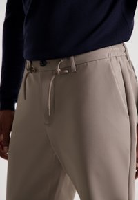 Pantaloni bej ajustați cu o croială relaxată, având închidere cu nasture, detalii cu șnur și textură netedă. Purtați cu un top de culoare bleumarin închis.