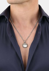 Collana a catena con pendente geometrico in argento, caratterizzata da una forma ottagonale, superficie lucida e bordi texturizzati; indossata sopra una maglietta scura.