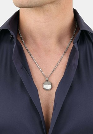 Zilveren geometrische hangerketting met een octagonale vorm, gepolijste oppervlakte en getextureerde randen; gedragen over een donkere blouse.