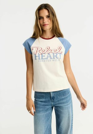 Młoda kobieta w białej i niebieskiej koszulce z napisem "Rebel Heart Forever Wild" oraz niebieskich dżinsach, stojąca na jednolitym białym tle.