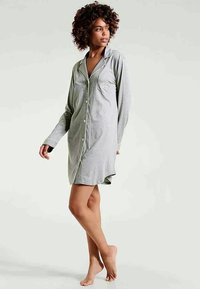 Nainen seisoo paljain jaloin, päällään pitkähihaisessa, nappivaatteisessa, vaaleanharmaassa yöpaidassa, jossa on valkoiset reunat ja pieni rintatasku.