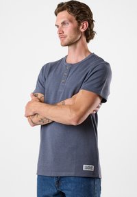 T-shirt grigia a maniche corte in stile Henley con texture a coste, tre bottoni in legno al collo e una piccola patch con logo sull'orlo.