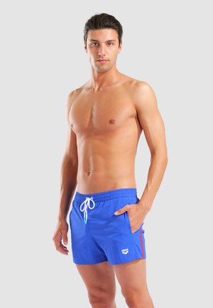 Skjorteløs mand står med den ene hånd i lommen iført blå badeshorts med hvid snøre og rød stribe på siden.