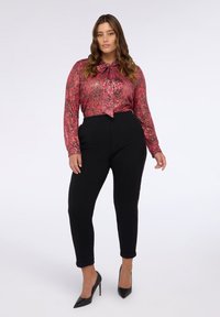 Blusa roja con estampado y mangas largas, con detalle de lazo, acompañada de pantalones ajustados negros y zapatos de tacón altos negros.