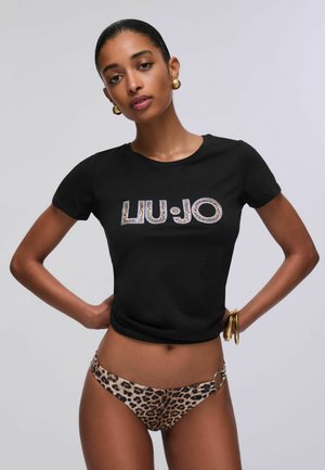 Mujer con camiseta negra corta con texto decorado "LIU•JO", braguitas de bikini con estampado de leopardo, pendientes de aro dorados y pulseras, de pie con las manos en las caderas.