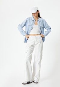 Camisa de botones azul claro sobre un top corto blanco, combinada con pantalones cargo blancos, una gorra blanca y zapatos grises con rayas negras.