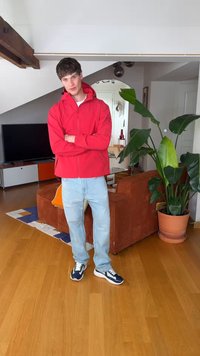 Νεαρός άνδρας με κόκκινο μπουφάν, ανοιχτόχρωμα τζιν και αθλητικά παπούτσια στέκεται με σταυρωμένα τα χέρια σε ένα σαλόνι με φυτά, τηλεόραση και πορτοκαλί καναπέ.