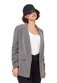Blazer grigio con design delle spalle strutturato, chiusura con un bottone e tasche davanti. Indossato sopra una maglietta bianca e pantaloni neri, abbinato a un cappellino nero.