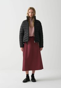 Zwarte gewatteerde jas met diagonale stiknaden, een hoge kraag, gecombineerd met een roze ritsvest en een bordeauxrode enkellange rok. Zwarte schoenen.