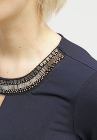 Marineblaues Oberteil mit einem mit Metallperlen und Verzierungen versehenen Be neckline, glattem Stoff und figurbetontem Schnitt.