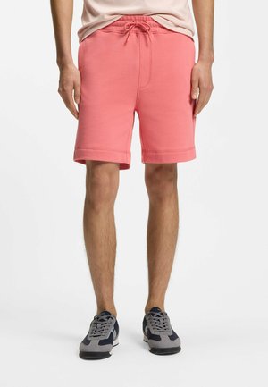 Mężczyzna ubrany w różowe szorty na sznurki oraz szare i granatowe sneakersy, stojący na jednolitym białym tle.