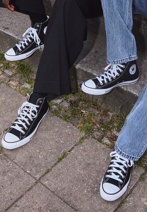 Black high-top canvas sneakers with white rubber soles and laces, featuring a circular logo on the side and paired with denim and black pants.