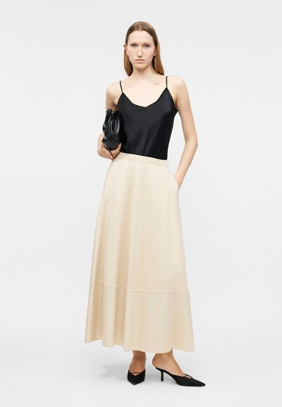 Γυναίκα που φοράει μαύρο αμάνικο τοπ, μακριά μπεζ φούστα, μαύρες μυτερές γόβες και κρατάει μαύρο τσάντα χειρός.