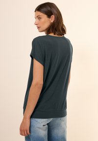 Maglietta corta in teal scuro, con un taglio morbido, scollatura rotonda e una texture morbida, abbinata a jeans in denim blu chiaro.