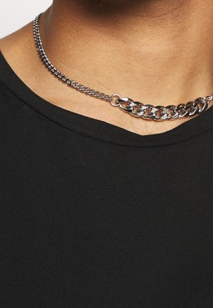 Gros plan sur un collier en chaîne métallique avec des maillons moyens et grands porté autour du cou d'une personne au-dessus d'une chemise noire.