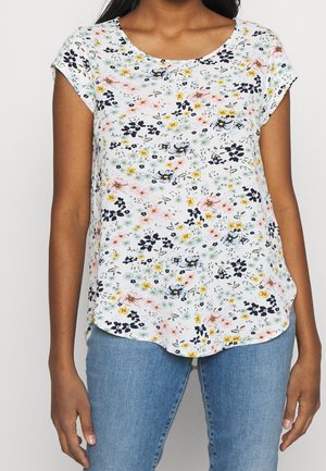 Vrouw met een wit topje met korte mouwen en een meerkleurige bloemenprint en een blauwe spijkerbroek, zichtbaar van schouders tot heupen.