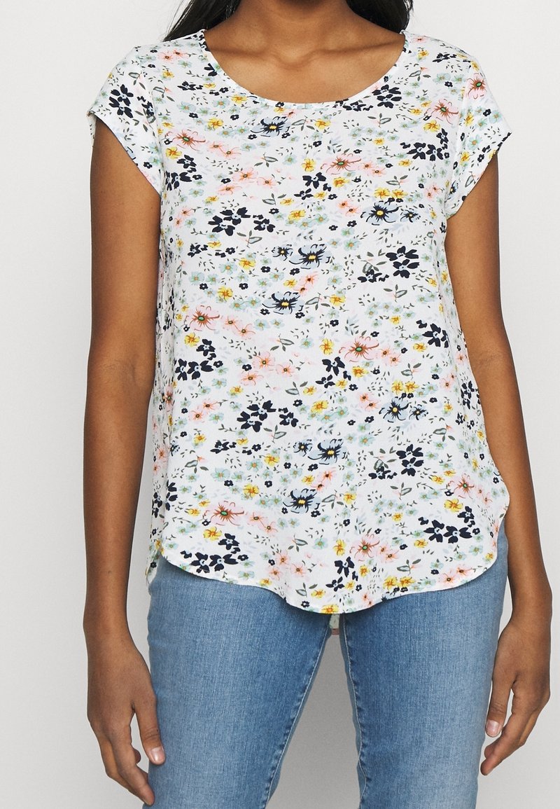 Femme portant un haut blanc à manches courtes avec un imprimé floral multicolore et un jean bleu, visible des épaules aux hanches.