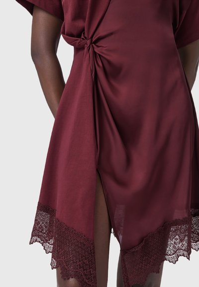 Robe en satin bordeaux avec des bordures en dentelle, détail de nœud sur le côté et fente haute sur le haut des cuisses, portée par une personne à la peau foncée au niveau du bas du torse et des jambes.