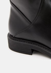 Stivale alla caviglia in pelle nera con una superficie liscia, leggero lucido e tacco impilato con cuciture visibili lungo la cucitura superiore.