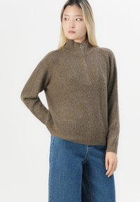 Maglione marrone a coste con collo alto, mezza cerniera e tasca frontale, abbinato a jeans a gamba larga blu chiaro.