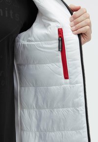 Interior de chaqueta blanca acolchada con un bolsillo rojo con cremallera y herrajes de cremallera negra. Tejido texturizado con patrones de costura horizontales.