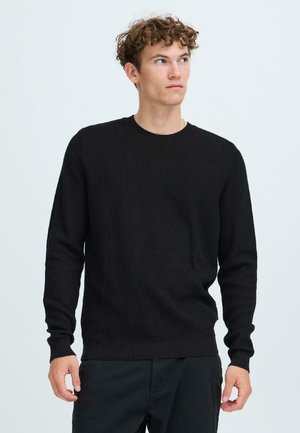 Svart strikket genser med en ribbestrikket tekstur, rund halsutskjæring og lange ermer, sammen med mørke bukser. Enkel, ensfarget design.