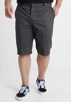 Homme portant un short gris foncé au genou et des baskets noires, bleues, blanches, debout les mains près des poches.