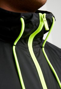 Svart jacka med hög krage, neongula dragkedjor och dragsko, med en slät yta och lättviktig design.