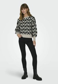 Maglione lavorato a maglia con un motivo zigzag nero e crema, colletto rotondo e maniche a palloncino. Abbinato a pantaloni skinny neri e stivali neri chunky.