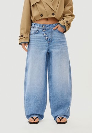 Kurz geschnittene beige Jacke mit Knopfdetails, kombiniert mit weitem, hoch tailliertem hellblauem Jeansstoff mit mehreren Knöpfen vorne und schwarzen Sandalen.