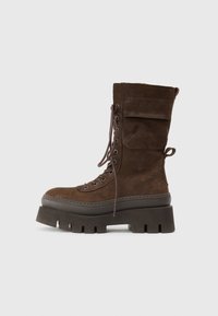 Stivali alla caviglia in suede marrone con lacci, dotati di una suola piatta chunky, tasca laterale, punta rotonda e occhielli in metallo per i lacci.
