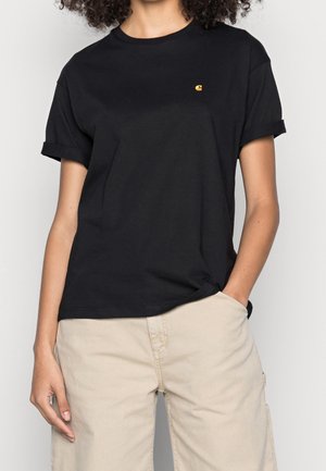 Persona con camiseta negra de mangas remangadas y un pequeño logo amarillo en el pecho izquierdo, y pantalones beige, con la mano en el bolsillo.