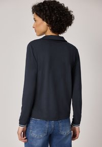 Femme aux cheveux bouclés portant une chemise noire à manches longues avec des rayures blanches sur les poignets et un jean bleu en denim, vue de dos sur un fond uni.