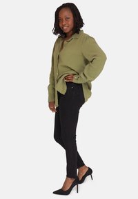 Camisa de botón texturizada verde olivo, jeans skinny negros y tacones altos negros con punta afilada. La modelo adopta una pose relajada.