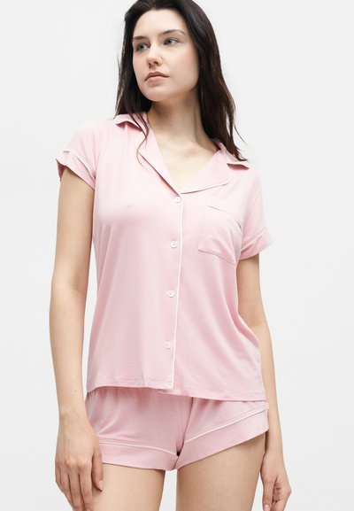 Νεαρή γυναίκα που φοράει ροζ ανοιχτόχρωμο κοντομάνικο πουκάμισο με κουμπιά και ταιριαστό σετ σορτς πιτζάμα, στέκεται μπροστά σε απλό λευκό φόντο.