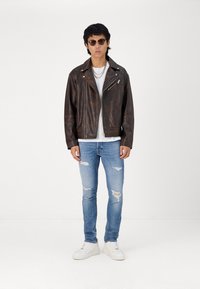 Giubbotto biker in pelle marrone con cerniere metallizzate, abbinato a una maglietta bianca, jeans blu strappati e sneakers bianche.