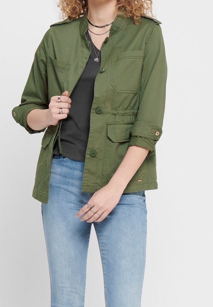 Femme portant une veste olive défaite aux manches retroussées sur un haut noir et un jean bleu clair, accessoirisée avec des bagues et des colliers superposés.