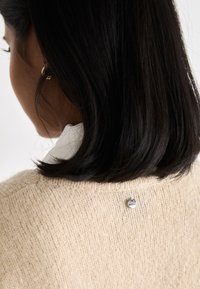 Femme aux cheveux noirs mi-longs portant un pull en tricot beige et un chemisier blanc, montrant un petit logo métallique circulaire au dos du pull.