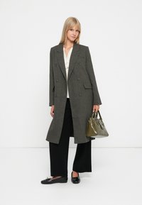 Dvojité sako s houndstooth vzorom v tmavozelenej a sivej farbe, doplnené krémovou blúzkou, čiernymi širokými nohavicami a zelenou kabelkou.