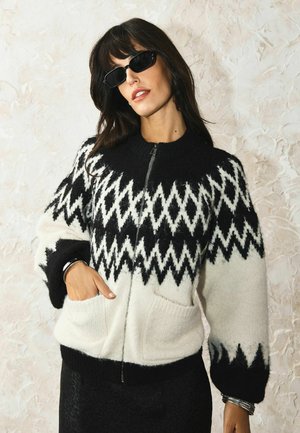 Vrouw met zwarte zonnebril en een zwart-wit zigzag patroon zip-up trui met zakken, poseert tegen een gestructureerde lichte muur.