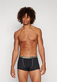 Jovem com cabelo cacheado vestindo cuecas boxer pretas com cós de marca, posando contra um fundo claro e simples.