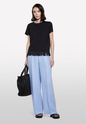 Schwarzes Kurzarmoberteil mit Fransendetail am Saum, kombiniert mit hellblauen weit geschnittenen Hosen. Schwarze strukturierte Tote-Tasche und dunkle Sandalen.