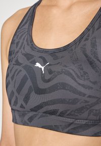 Puma sportski grudnjak u tamno sivoj boji, s teksturiranim uzorkom u obliku valova i bijelim logotipom. Izrađen od rastezljivog, prozračnog materijala. Podesive naramenice.