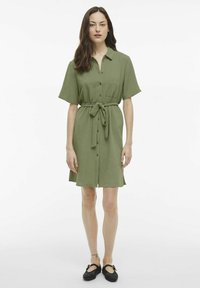 Šviesiai žalios spalvos trumpų rankovių suknelė, pasižyminti tekstūruota medžiaga, mygtukų užsegimu, dviem krūtinės kišenėmis ir diržu liemeniui.
