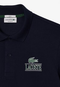 Polo navy de tela texturizada con un logo de cocodrilo bordado en verde y "LACOSTE" en blanco debajo del logo. Corte regular.