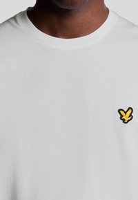 Mørkhyet person iført en almindelig hvid T-shirt med rund hals og et lille gult fuglelogo på venstre bryst.