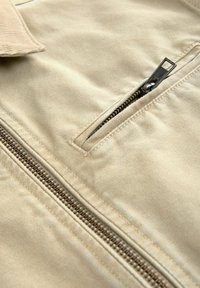 Vaalean beige takki, jossa on edessä vetoketjullinen tasku mustalla vetoketjulla, teksturoitu kangas ja näkyvät ompeluyksityiskohdat. Tasainen pinta.
