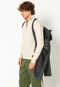 Lichtbeige ritsvest, groene corduroybroek en een donkergroene pufferjas over de schouder, met zwarte accenten en labels.