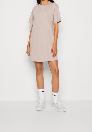 Oversized, short-sleeve dress in light beige cotton. Features a small white Nike logo on the chest. Paired with white socks and light gray sneakers.
