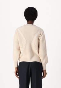 Maglione crema a trecce con collo rotondo, motivi testurizzati e polsini a coste. Il retro presenta un design geometrico.